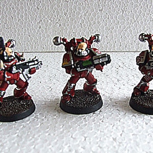 Puede incluir: Tres figuras en miniatura de soldados blindados rojos y plateados, cada uno sosteniendo un arma. Las figuras están sobre bases oscuras y circulares. El fondo es una superficie blanca texturizada.