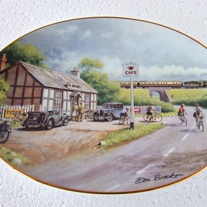 May include: A vintage oval ceramic plate featuring a painted scene of a charming English countryside cafe. The cafe is a white building with a brown roof and a sign that reads "The Old Inn". A car is parked in front of the cafe, and a train is passing by in the background. The plate is signed "Don Bracken" in the bottom right corner.