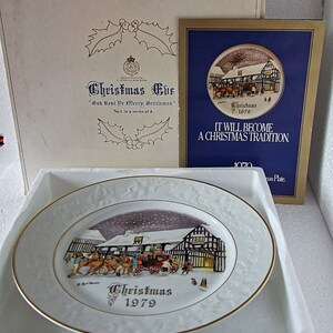 May include: A white porcelain Christmas plate with a gold rim. The plate features a winter scene with a building, horses, and people. The text "Christmas 1979" is printed on the plate.