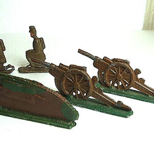 May include: A collection of vintage toy military figures and artillery. The set includes two cannons, a tank, and two kneeling soldiers. The toys are painted in shades of brown, green, and bronze.