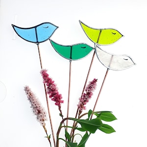 May include: Four colourful glass bird garden stakes with copper wire stems. The birds are blue, green, yellow, and white. The stakes are arranged in a bouquet with pink flowers and green leaves.