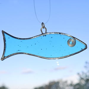 Pu&ograve; includere: Un ornamento a forma di pesce in vetro colorato azzurro, con un contorno in metallo color argento e un ricciolo decorativo. Il pesce &egrave; sospeso da un filo sottile e trasparente su uno sfondo di cielo blu.