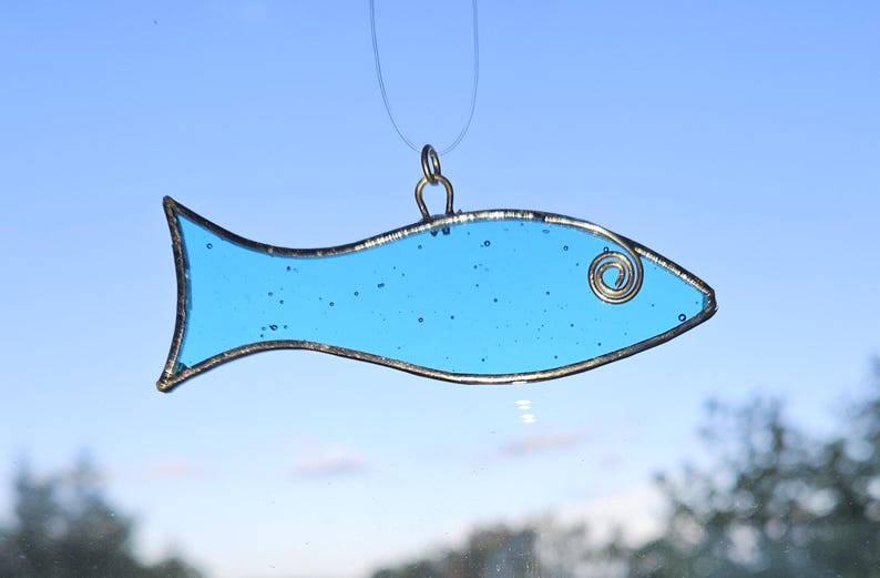 Pu&ograve; includere: Un ornamento a forma di pesce in vetro colorato azzurro, con un contorno in metallo color argento e un ricciolo decorativo. Il pesce &egrave; sospeso da un filo sottile e trasparente su uno sfondo di cielo blu.