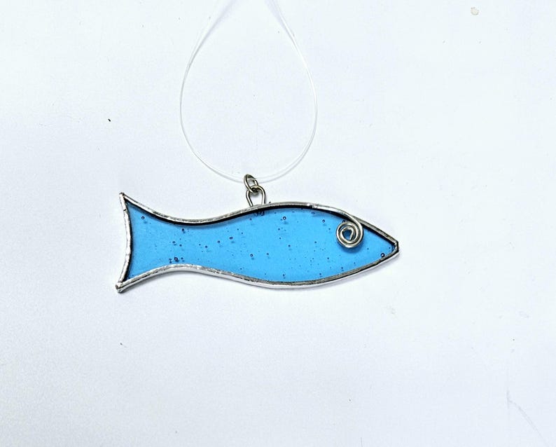 Pu&ograve; includere: Ciondolo a forma di pesce in vetro colorato, con corpo azzurro chiaro e contorno in metallo argentato. Il pesce ha un occhio a spirale ed &egrave; sospeso da un cordino trasparente. Il ciondolo misura circa 7.6 cm di lunghezza.