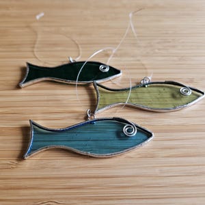 Pu&ograve; includere: Tre ornamenti a forma di pesce in vetro colorato nelle tonalit&agrave; del verde e del blu, ciascuno con un contorno in metallo color argento e un piccolo anello per appenderli. I pesci sono disposti su una superficie di legno marrone chiaro.