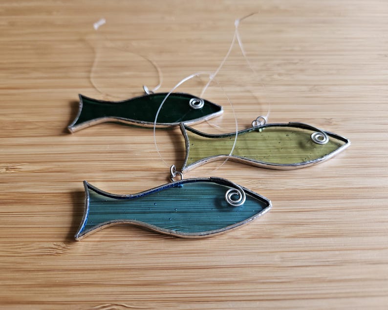 Pu&ograve; includere: Tre ornamenti a forma di pesce in vetro colorato nelle tonalit&agrave; del verde e del blu, ciascuno con un contorno in metallo color argento e un piccolo anello per appenderli. I pesci sono disposti su una superficie di legno marrone chiaro.