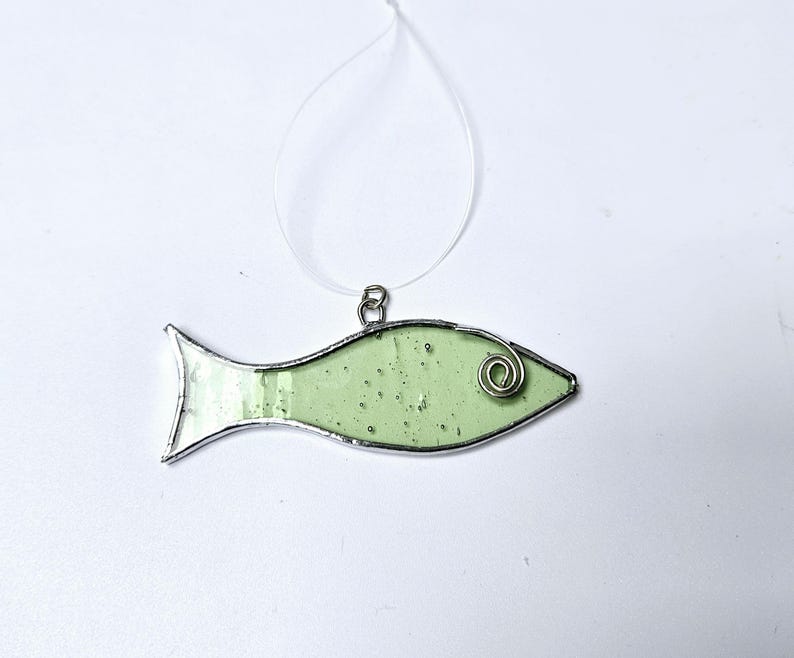 Pu&ograve; includere: Un ornamento a forma di pesce in vetro colorato verde chiaro, con un contorno in metallo color argento e un dettaglio a spirale in filo di ferro. Il pesce &egrave; sospeso da un filo trasparente, adatto per essere appeso. Un oggetto decorativo.