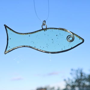 Pu&ograve; includere: Un acchiappasole a forma di pesce in vetro colorato azzurro con un contorno in metallo color oro e un dettaglio a spirale in filo di ferro. Il pesce &egrave; sospeso da una corda trasparente su uno sfondo di cielo blu. Decorazione.