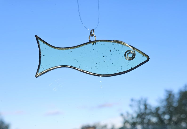 Pu&ograve; includere: Un acchiappasole a forma di pesce in vetro colorato azzurro con un contorno in metallo color oro e un dettaglio a spirale in filo di ferro. Il pesce &egrave; sospeso da una corda trasparente su uno sfondo di cielo blu. Decorazione.