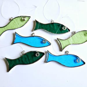 Pu&ograve; includere: Una collezione di sei ornamenti a forma di pesce in vetro colorato, nei toni del verde e del blu. Ogni pesce ha un occhio di filo d'argento e un anello d'argento per appenderlo. I pesci sono disposti su uno sfondo bianco.