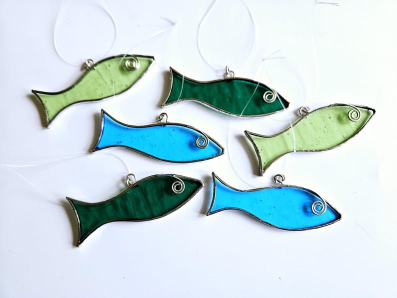 Pu&ograve; includere: Una collezione di sei ornamenti a forma di pesce in vetro colorato, nei toni del verde e del blu. Ogni pesce ha un occhio di filo d'argento e un anello d'argento per appenderlo. I pesci sono disposti su uno sfondo bianco.