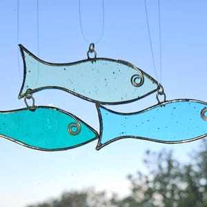 Pu&ograve; includere: Tre pesci in vetro colorato blu e turchese sono appesi a fili sottili. Ogni pesce ha un contorno di filo e un ricciolo decorativo. Lo sfondo &egrave; un cielo azzurro e alcuni rami di alberi.