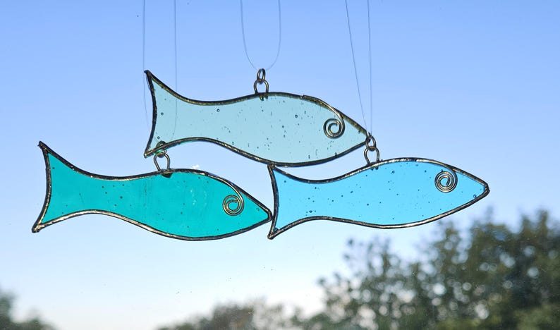 Pu&ograve; includere: Tre pesci in vetro colorato blu e turchese sono appesi a fili sottili. Ogni pesce ha un contorno di filo e un ricciolo decorativo. Lo sfondo &egrave; un cielo azzurro e alcuni rami di alberi.