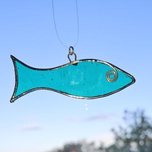 Pu&ograve; includere: Un ornamento a forma di pesce in vetro colorato turchese con un contorno in filo dorato e un dettaglio a spirale per l'occhio. Il pesce &egrave; sospeso da un filo sottile e trasparente, su uno sfondo di cielo blu. Un oggetto decorativo.