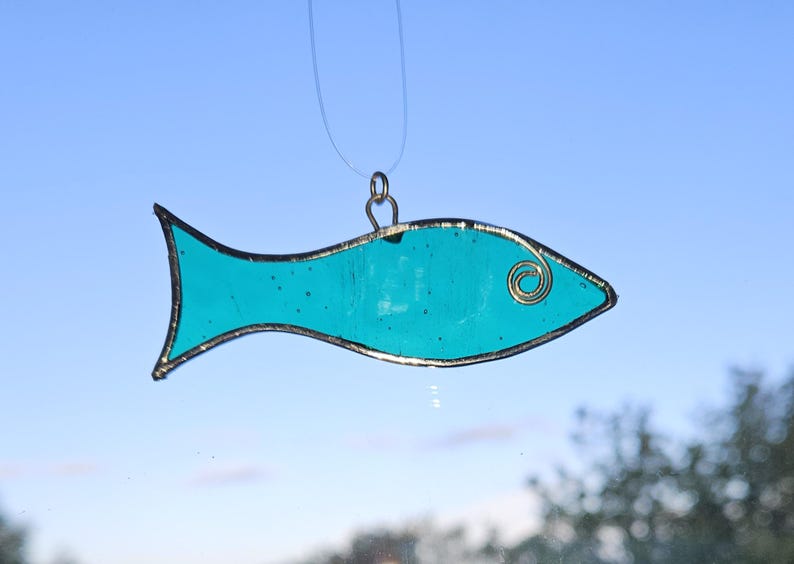 Pu&ograve; includere: Un ornamento a forma di pesce in vetro colorato turchese con un contorno in filo dorato e un dettaglio a spirale per l'occhio. Il pesce &egrave; sospeso da un filo sottile e trasparente, su uno sfondo di cielo blu. Un oggetto decorativo.