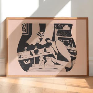 May include: A framed art print featuring a black and white illustration of a person working at a pottery wheel. The artwork depicts a studio scene with a dog, shelves of pottery, and various tools. The frame is made of light brown wood.
