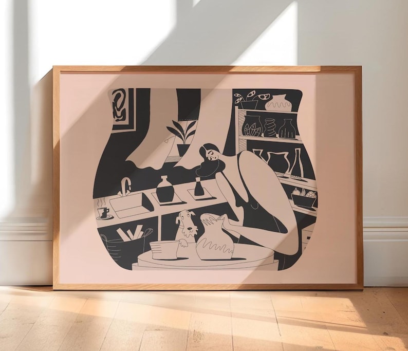 May include: A framed art print featuring a black and white illustration of a person working at a pottery wheel. The artwork depicts a studio scene with a dog, shelves of pottery, and various tools. The frame is made of light brown wood.