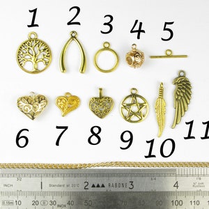 Op de afbeelding: Elf goudkleurige metalen bedels met verschillende ontwerpen, waaronder een boom, een wensbeen, een cirkel, een bal, een staaf, een hart, een veer en een vleugel. De bedels zijn genummerd van 1 tot en met 11. Een gouden ketting en een liniaal zijn ook zichtbaar op de afbeelding.