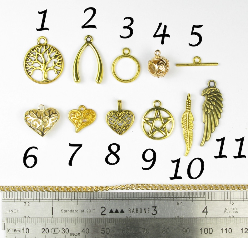 Op de afbeelding: Elf goudkleurige metalen bedels met verschillende ontwerpen, waaronder een boom, een wensbeen, een cirkel, een bal, een staaf, een hart, een veer en een vleugel. De bedels zijn genummerd van 1 tot en met 11. Een gouden ketting en een liniaal zijn ook zichtbaar op de afbeelding.