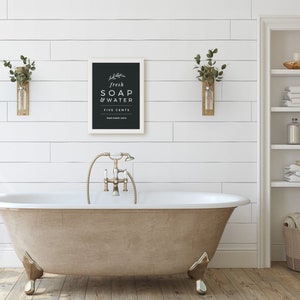 May include: A white framed sign with black lettering that reads "fresh soap & water five cents" hanging on a white shiplap wall above a freestanding bathtub with gold fixtures.