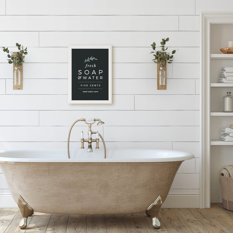 May include: A white framed sign with black lettering that reads "fresh soap & water five cents" hanging on a white shiplap wall above a freestanding bathtub with gold fixtures.