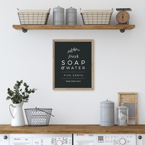 May include: Black and white sign with the text "fresh SOAP & WATER FIVE CENTS hand towels extra" hanging on a wooden shelf in a laundry room. The shelf is decorated with metal containers and baskets.