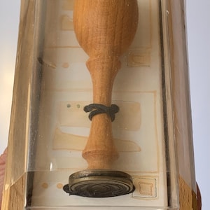 May include: A wooden stamp with a round handle and a metal base, displayed in a clear plastic case with a wooden frame. The stamp is in a box with a red circle logo.