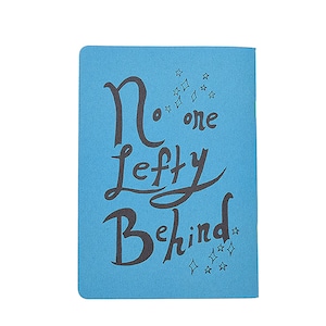 Puede incluir: Una libreta azul con el texto "No one left behind" en letras cursivas negras. Hay pequeñas estrellas alrededor del texto.