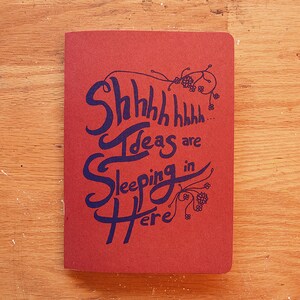 Puede incluir: Un cuaderno rojo con las palabras "Shhhh Ideas are Sleeping in Here" en escritura azul oscuro. El cuaderno est&aacute; sobre una superficie de madera.