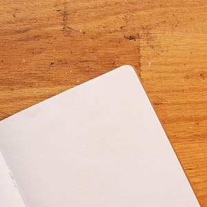 Puede incluir: Cuaderno abierto con p&aacute;ginas blancas. El cuaderno est&aacute; abierto en una doble p&aacute;gina, con las p&aacute;ginas en &aacute;ngulo hacia la esquina superior derecha. El fondo es una superficie de madera con un tono marr&oacute;n c&aacute;lido.