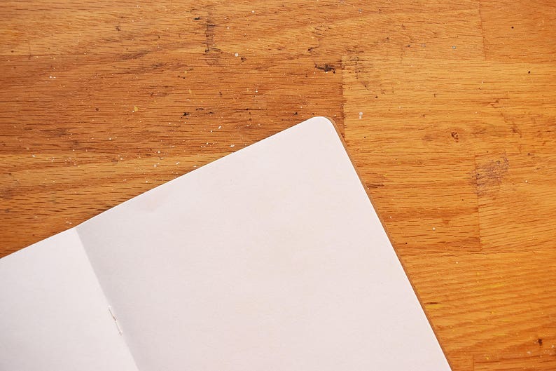 Puede incluir: Cuaderno abierto con p&aacute;ginas blancas. El cuaderno est&aacute; abierto en una doble p&aacute;gina, con las p&aacute;ginas en &aacute;ngulo hacia la esquina superior derecha. El fondo es una superficie de madera con un tono marr&oacute;n c&aacute;lido.