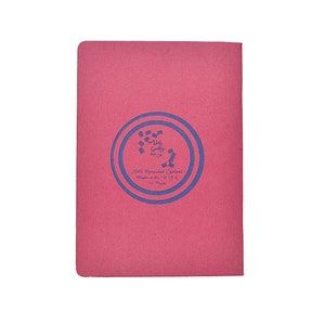 Puede incluir: Un cuaderno rosa con un dise&ntilde;o circular azul y texto. El texto dice "100% Recycled Content Made in the U.S.A. 12 Pages". El cuaderno tiene una forma rectangular simple.