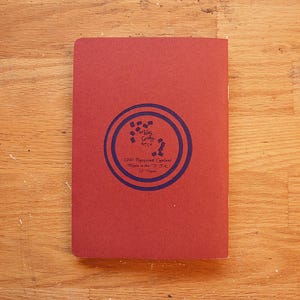 Puede incluir: Un cuaderno rojo rectangular cerrado con un dise&ntilde;o circular azul en la portada. El dise&ntilde;o incluye texto y un patr&oacute;n. El texto dice "100% Recycled Content Made in the U.S.A." en la portada.
