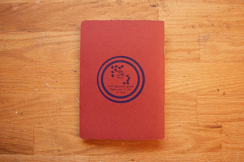 Puede incluir: Un cuaderno rojo rectangular cerrado con un dise&ntilde;o circular azul en la portada. El dise&ntilde;o incluye texto y un patr&oacute;n. El texto dice "100% Recycled Content Made in the U.S.A." en la portada.