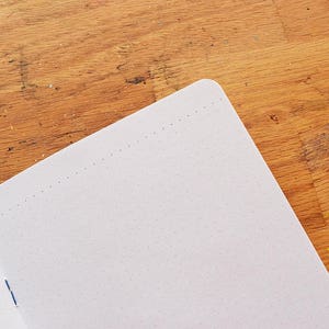 May include: A white notebook with a dotted grid pattern and a blue binding. The notebook is open and lying on a wooden surface.