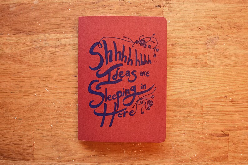 Puede incluir: Un cuaderno rojo con las palabras "Shhhh Ideas are Sleeping in Here" en escritura azul oscuro. El cuaderno est&aacute; sobre una superficie de madera.