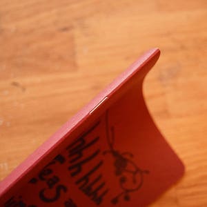 Puede incluir: Una funda de tel&eacute;fono roja con un dise&ntilde;o curvo. La funda tiene un gr&aacute;fico negro y texto en la superficie. La funda est&aacute; sujeta por una mano y el fondo es una superficie de madera.