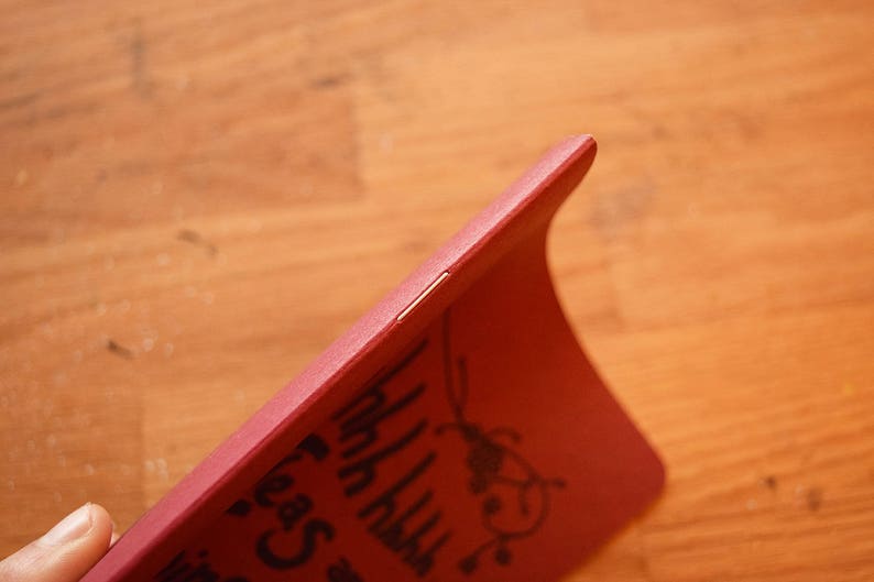 Puede incluir: Una funda de tel&eacute;fono roja con un dise&ntilde;o curvo. La funda tiene un gr&aacute;fico negro y texto en la superficie. La funda est&aacute; sujeta por una mano y el fondo es una superficie de madera.