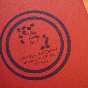 Puede incluir: Primer plano de un cuaderno rojo con un dise&ntilde;o circular azul oscuro. El dise&ntilde;o incluye el texto "The Witty Gritty Paper Co." y "1001 Recycled Content Made in the U.S.A. 32 Pages."