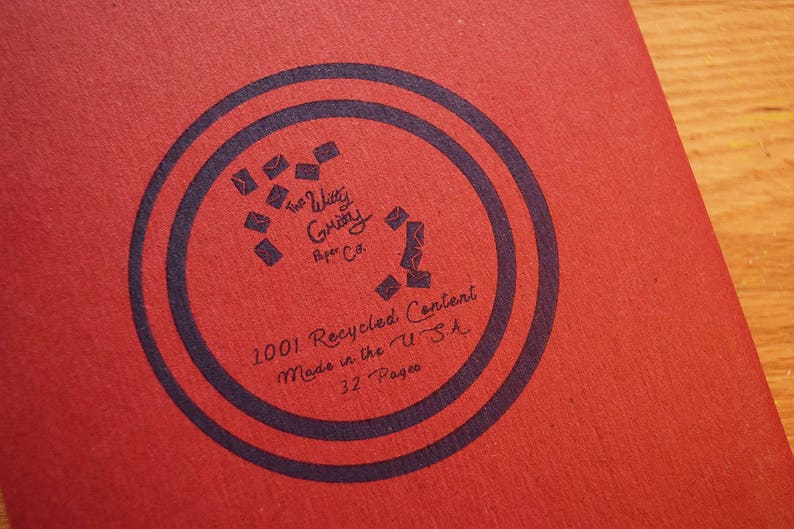 Puede incluir: Primer plano de un cuaderno rojo con un dise&ntilde;o circular azul oscuro. El dise&ntilde;o incluye el texto "The Witty Gritty Paper Co." y "1001 Recycled Content Made in the U.S.A. 32 Pages."