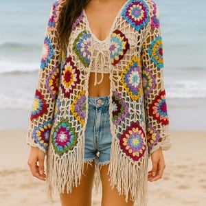 Puede incluir: Un cárdigan de crochet beige con frente abierto y patrones florales coloridos. La prenda de manga larga presenta un cuello en V y flecos a lo largo del dobladillo inferior. Los diseños florales incorporan tonos de rosa, azul, amarillo y morado.