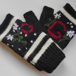 May include: A pair of black fingerless gloves featuring white striped cuffs and embroidered floral details. One glove displays a red letter C and the other a red letter G, both surrounded by small white flowers with green stems and white dots.