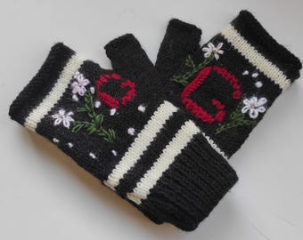 Puede incluir: Un par de guantes negros sin dedos con puños de rayas blancas y detalles florales bordados. Un guante muestra una letra C roja y el otro una letra G roja, ambos rodeados de pequeñas flores blancas con tallos verdes y puntos blancos.