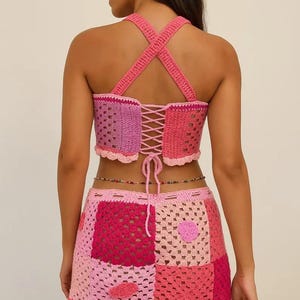 May include: A two-piece crochet set featuring a pink and magenta patchwork crop top with a lace-up back and crisscross straps, paired with a matching mini skirt. The set is crafted from various shades of pink yarn in a square-based pattern.