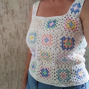Pode incluir: Um top branco de crochê com decote quadrado. O top é feito com quadrados de vovó em cores pastel, incluindo rosa, azul, amarelo e verde.