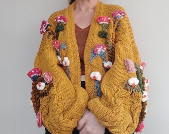 Pode incluir: Um cardigan de malha amarelo mostarda com um padrão de malha trançada e enfeites 3D de flores e cogumelos.