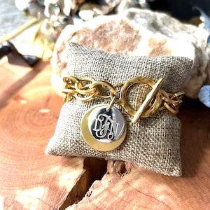 May include: A gold-toned chain bracelet featuring a toggle clasp and two circular charms. One charm is a solid gold-toned disc, while the smaller silver-toned disc layered on top is engraved with the stylized monogram letters DJN.