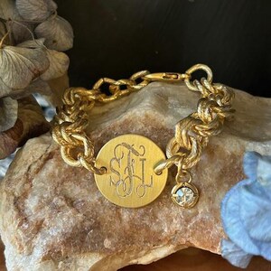 May include: A gold chain bracelet with a round charm engraved with the initials "SJL" and a small crystal charm.
