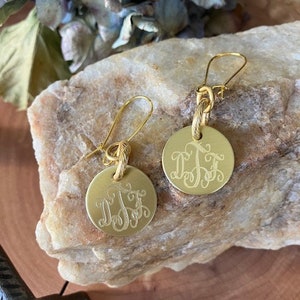 May include: A pair of gold-toned earrings with a round, engraved monogram charm. The charms are suspended from a twisted gold wire and have a hook closure.
