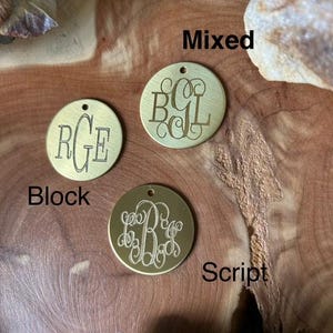 May include: Three brass discs with different monogram styles: "RGE" in block letters, "BCL" in mixed script, and "B" in a cursive script. The discs are on a wooden surface with the words "Block", "Mixed", and "Script" written below each disc.
